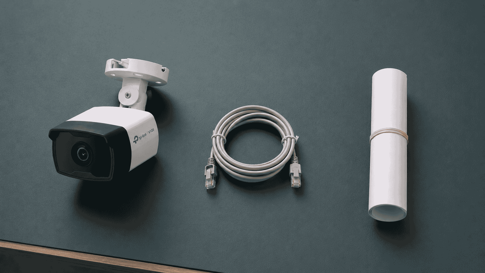 VIGI camera and its metal mounting bracket laid out on a workbench beside a coil of network cable and a rolled site plan